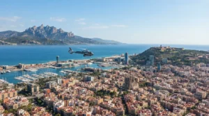 A helicopter flies over the Barcelona coastline, port, and city landmarks on a bright, sunny day.