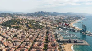 A red and white helicopter flies over the sunny Barcelona coastline and harbor against a mountainous backdrop.