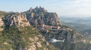 A small white helicopter in the air with the jagged rock pinnacles of Montserrat and its monastery on a ledge behind it
