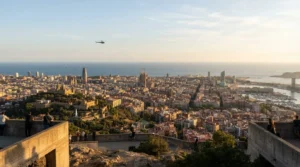 Barcelona rooftops and the sea seen from a hilltop viewpoint in warm light, with a small helicopter far off in the sky