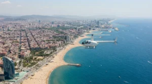Small white helicopter flying over Barceloneta beach and the Barcelona waterfront, the coast stretching north to the horizon