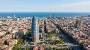 Aerial daylight view of the rounded Torre Glories tower above Barcelona's street grid, with the sea on the horizon