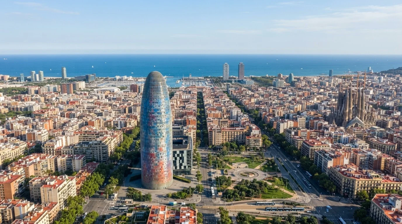 Aerial daylight view of the rounded Torre Glories tower above Barcelona's street grid, with the sea on the horizon