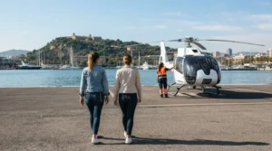 Two passengers in light jackets and flat shoes walk with ground crew to a white helicopter on a Port Vell helipad